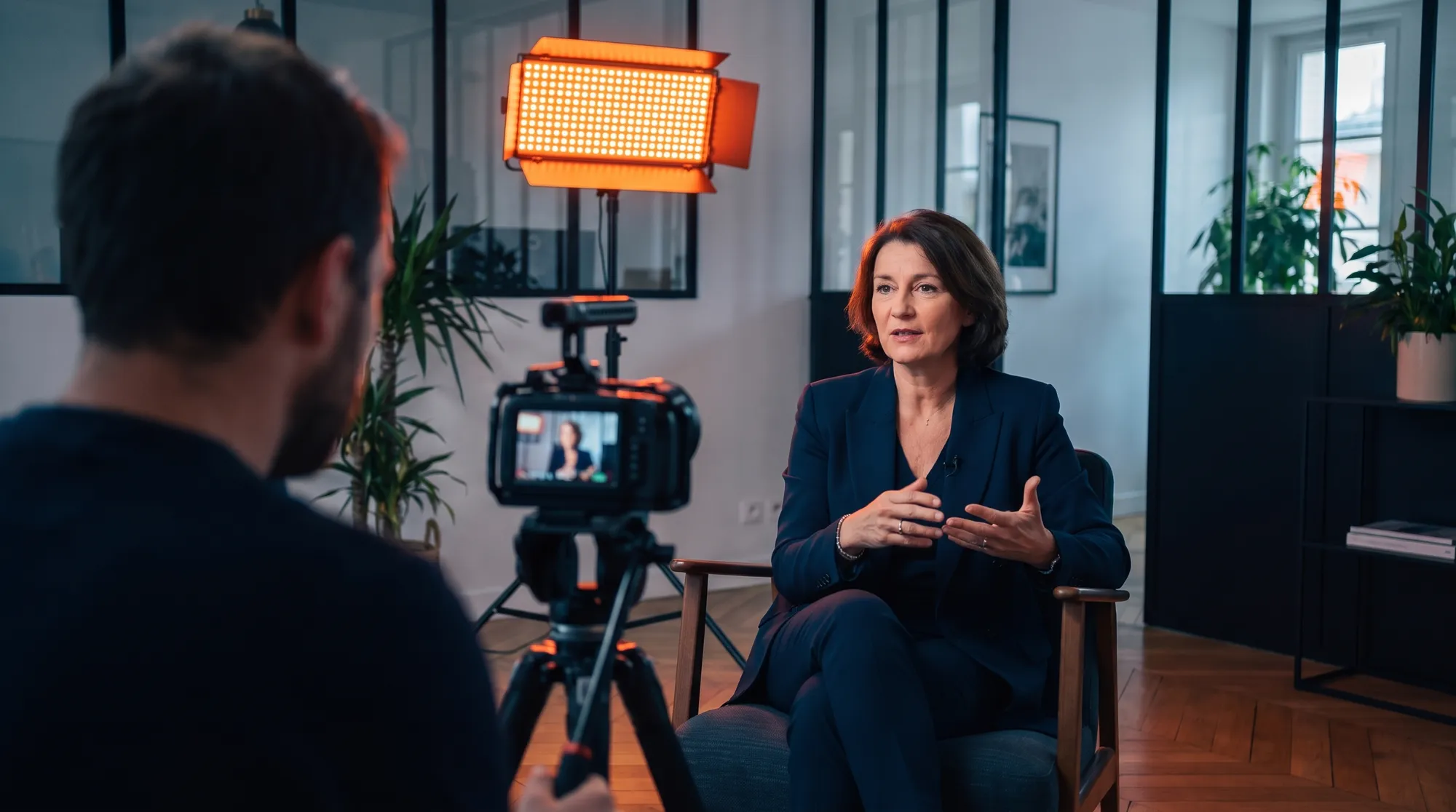 Dirigeante interviewée dans des bureaux à verrières, filmée par un cadreur sur trépied, panneau LED orange en contre-jour
