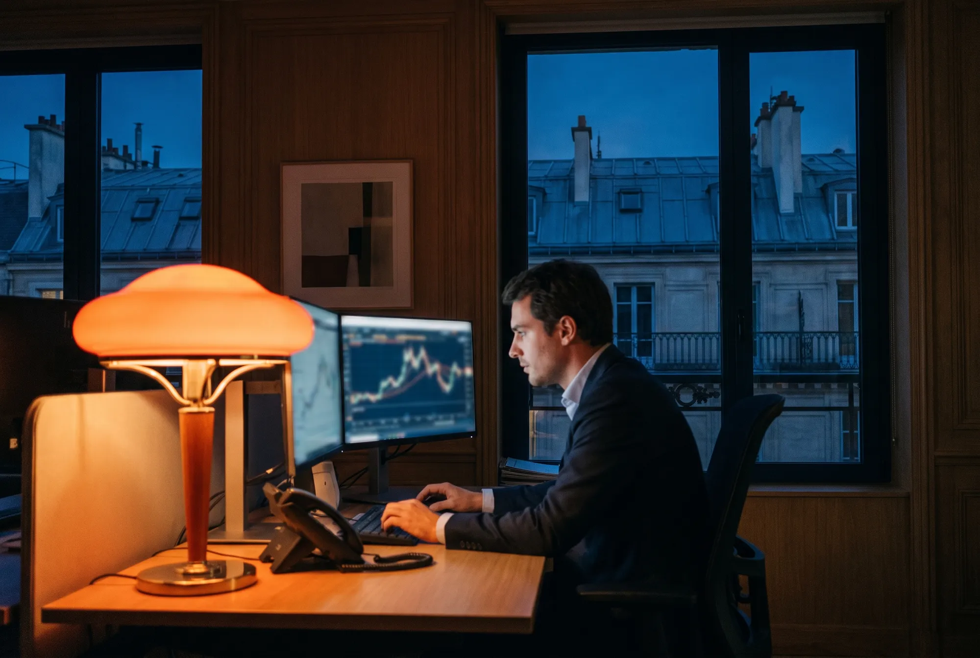 Analyste d’une banque travaillant le soir devant deux écrans de courbes, lampe orangée et toits de zinc parisiens aux fenêtres