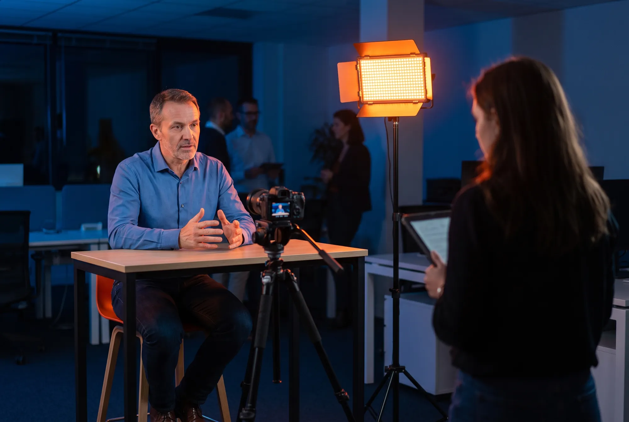 Formateur interne en chemise bleue expliquant un procédé face caméra dans un open space, pendant le tournage d’un module e-learning