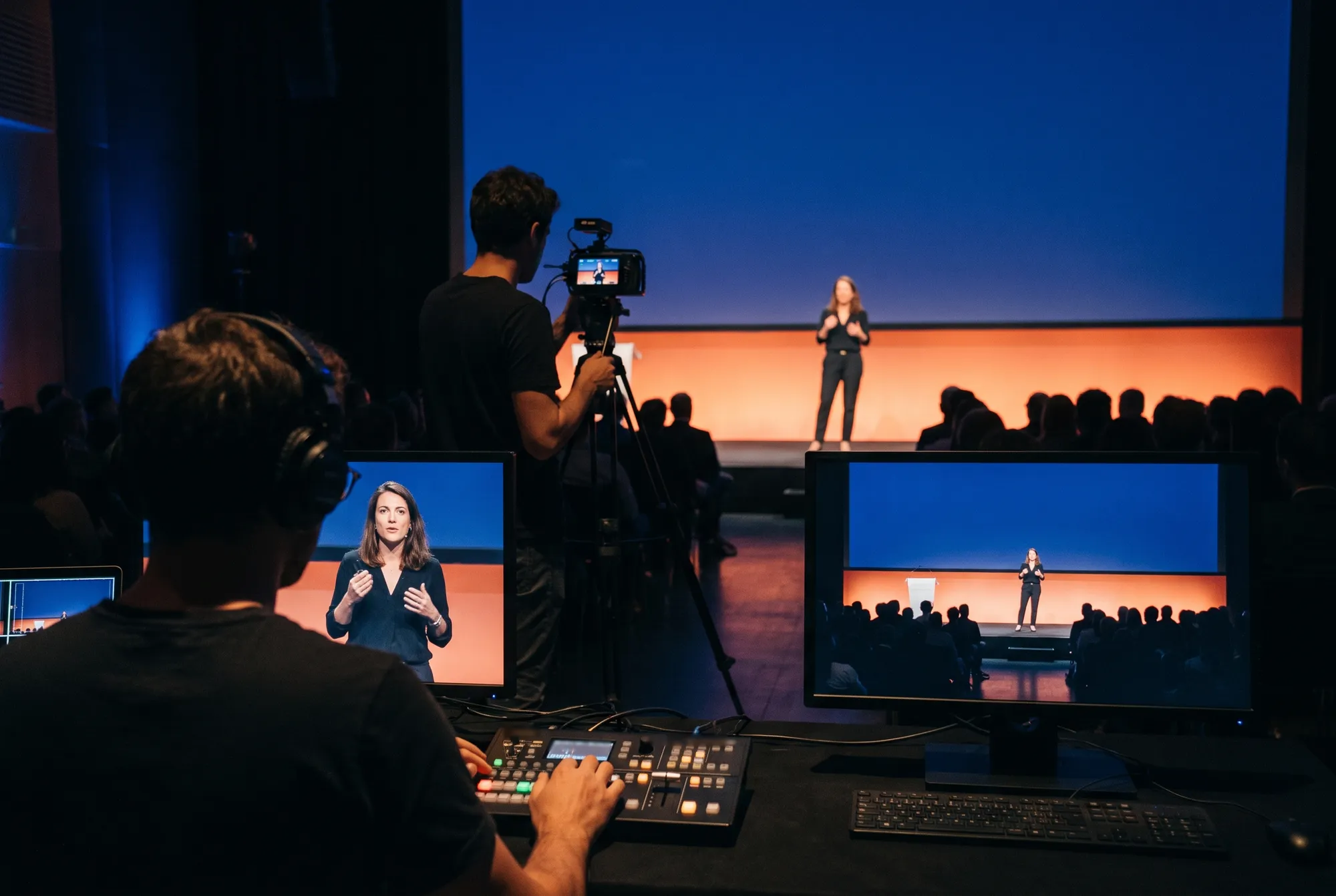 Régie de captation multicaméra d’une plénière, un technicien au mélangeur vidéo et l’intervenante sur scène au loin