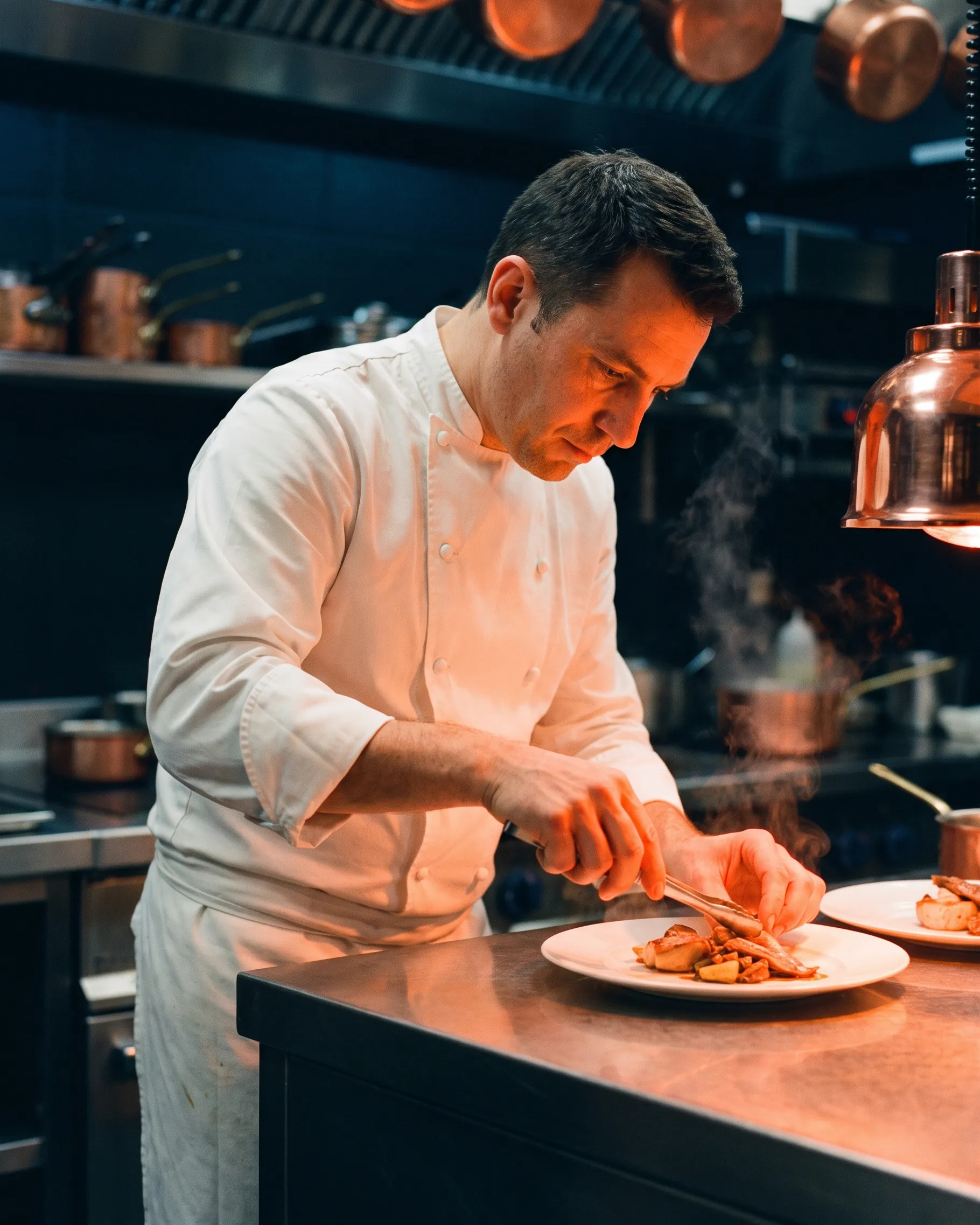 Chef en veste blanche dressant une assiette à la pince sur le passe d’une cuisine d’hôtel, sous une lampe chauffante